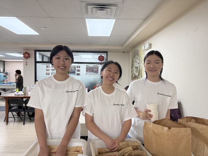 Nourish & Knowledge volunteers standing behind bins of packed meals, holding cups of soup.
